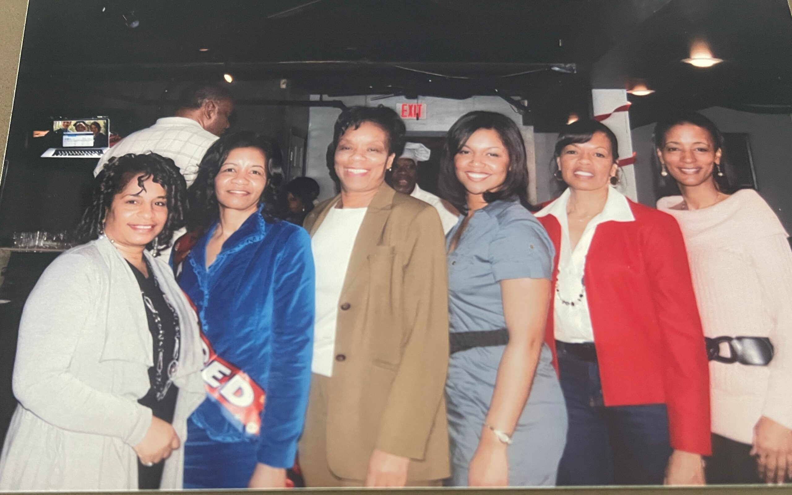 Six women posing together for a group photo at an indoor social event, all smiling at the camera.