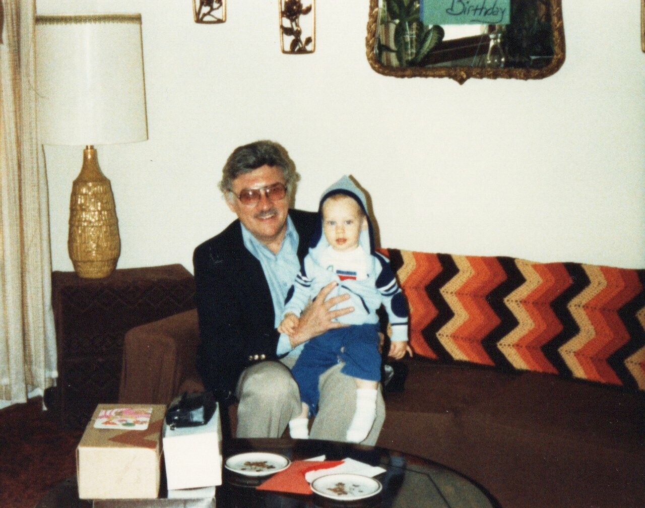 Grandfather in a blazer holding a baby on a patterned sofa in a vintage living room, with a lamp and retro decor nearby.