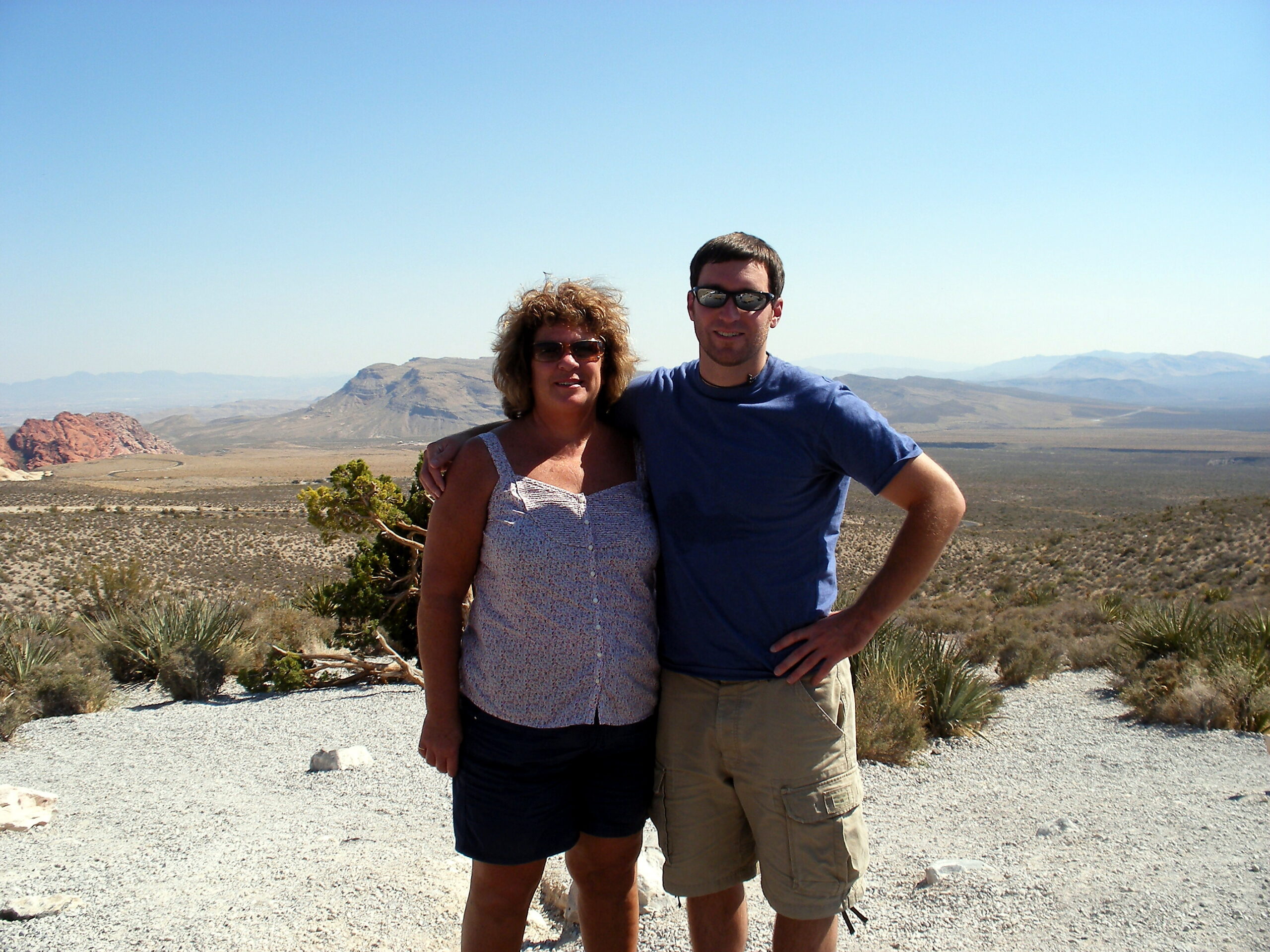 Two people standing arm-in-arm in a desert landscape with mountains and a clear blue sky.