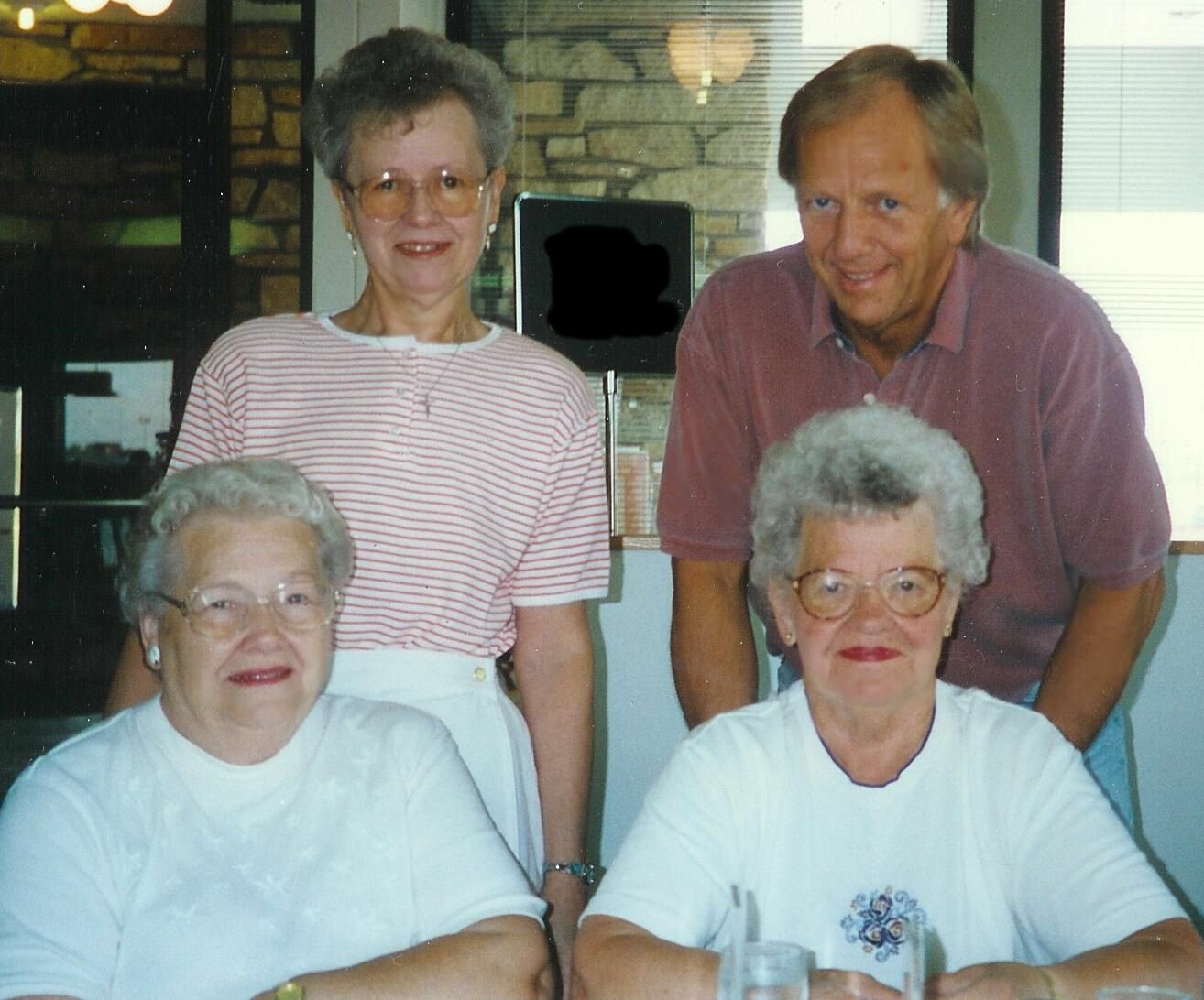 Four adults posing for a photo, two seated at a table in front and two standing behind, all smiling indoors.