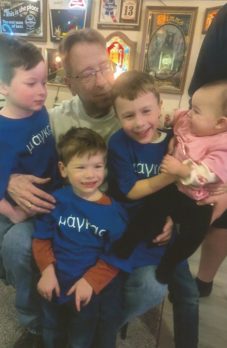 Father with glasses sits among four children: three boys in blue shirts and a baby girl in pink, all smiling in a room full of framed memorabilia behind them.