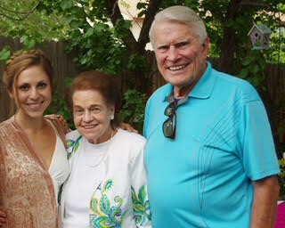 Three people posing outdoors and smiling: a middle-aged woman on the left, an elderly woman in the center, and an older man on the right wearing a teal polo and sunglasses hanging from his shirt.
