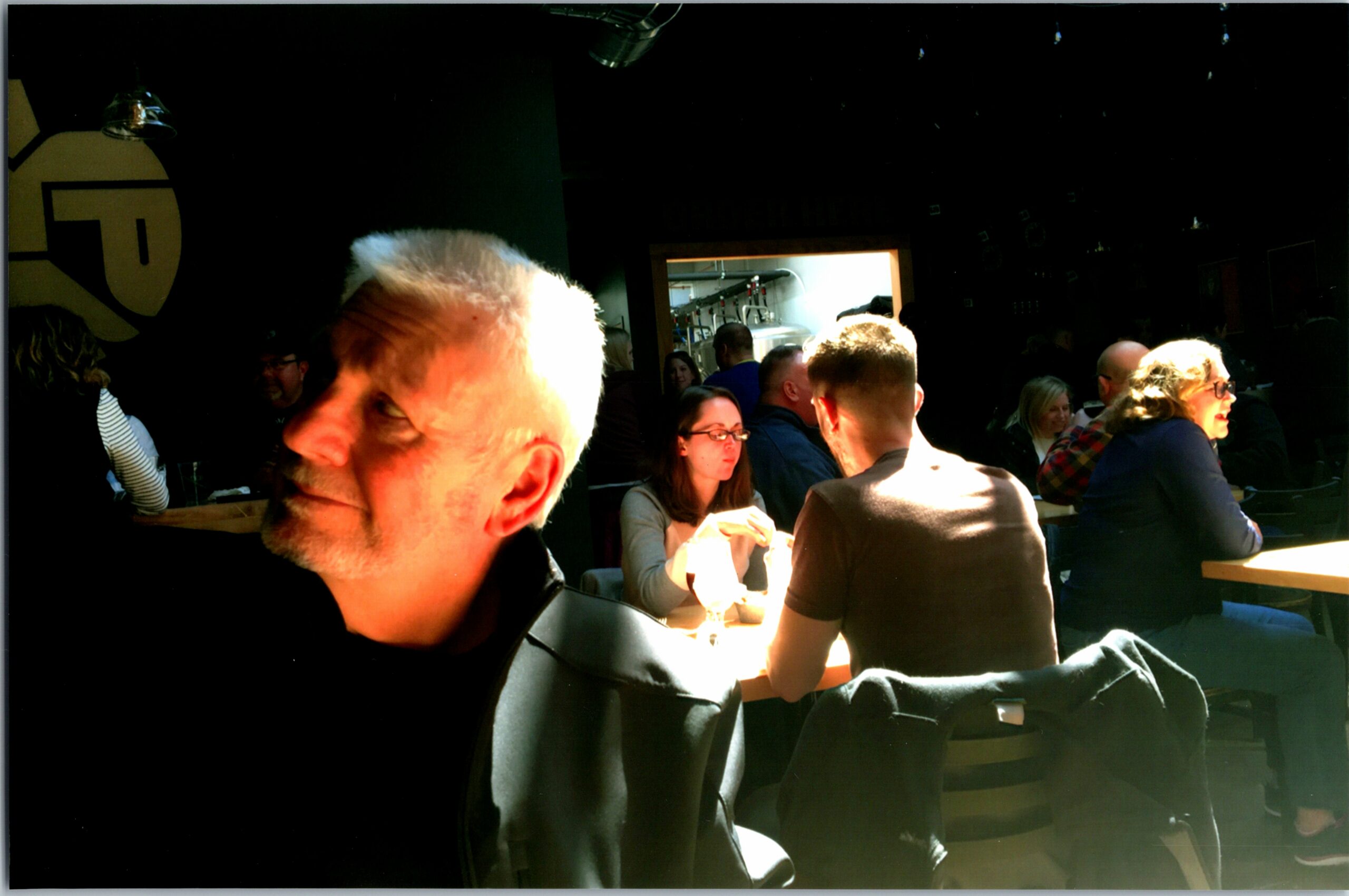 Crowded indoor dining area with people at tables; a man with white hair is in the foreground, sunlit.