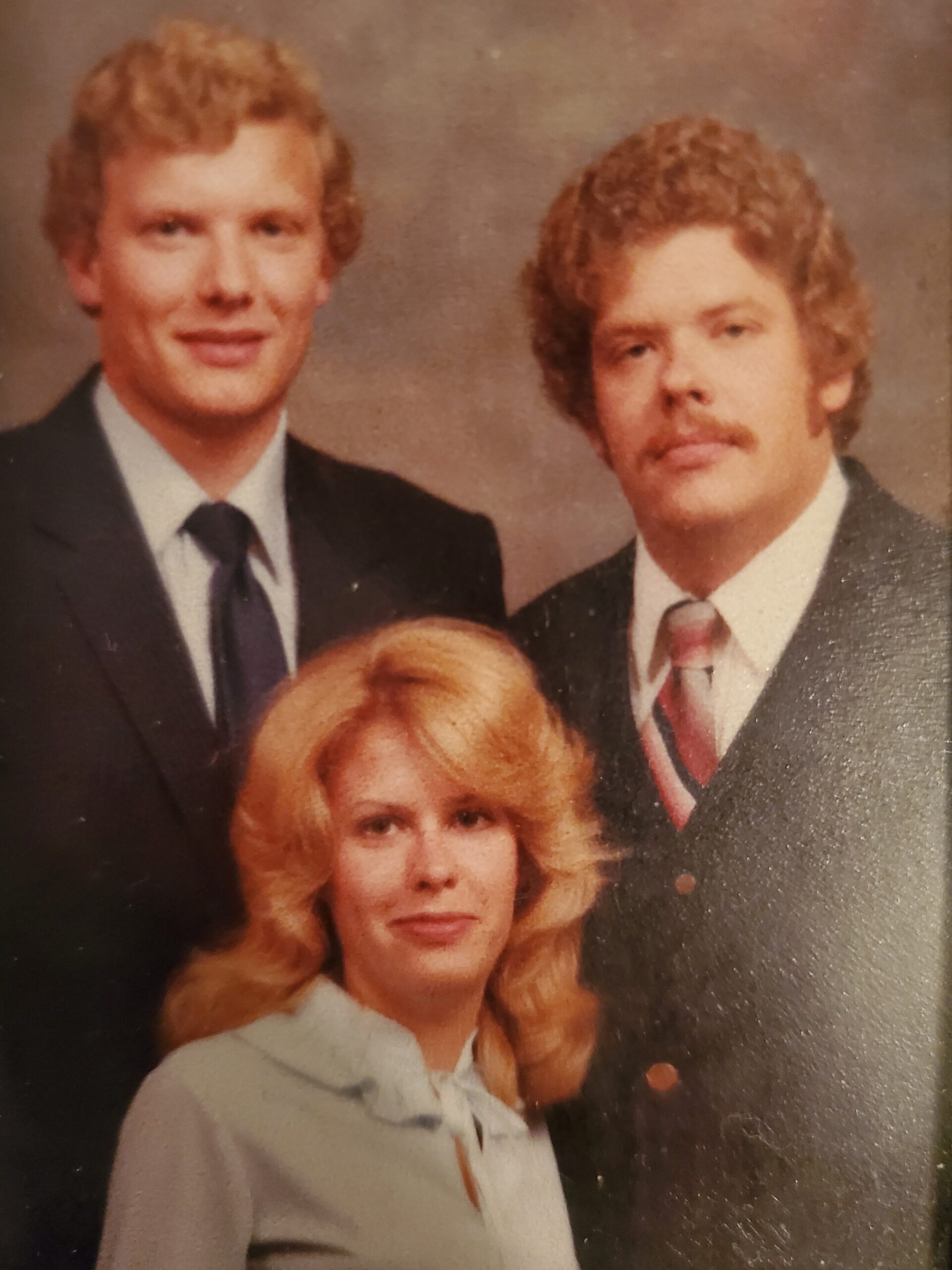 Three people pose for a formal portrait: a blonde woman in a light blazer in front, with two men in suits behind her.