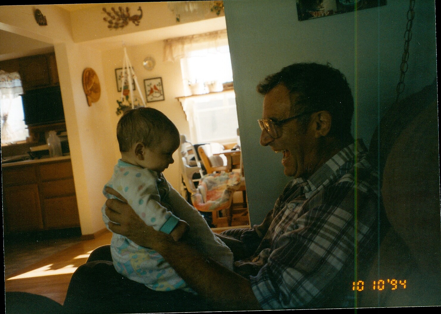Man in plaid shirt smiling while holding a baby facing him in a cozy home living room. Warm light, kitchen and dining area visible in the background.