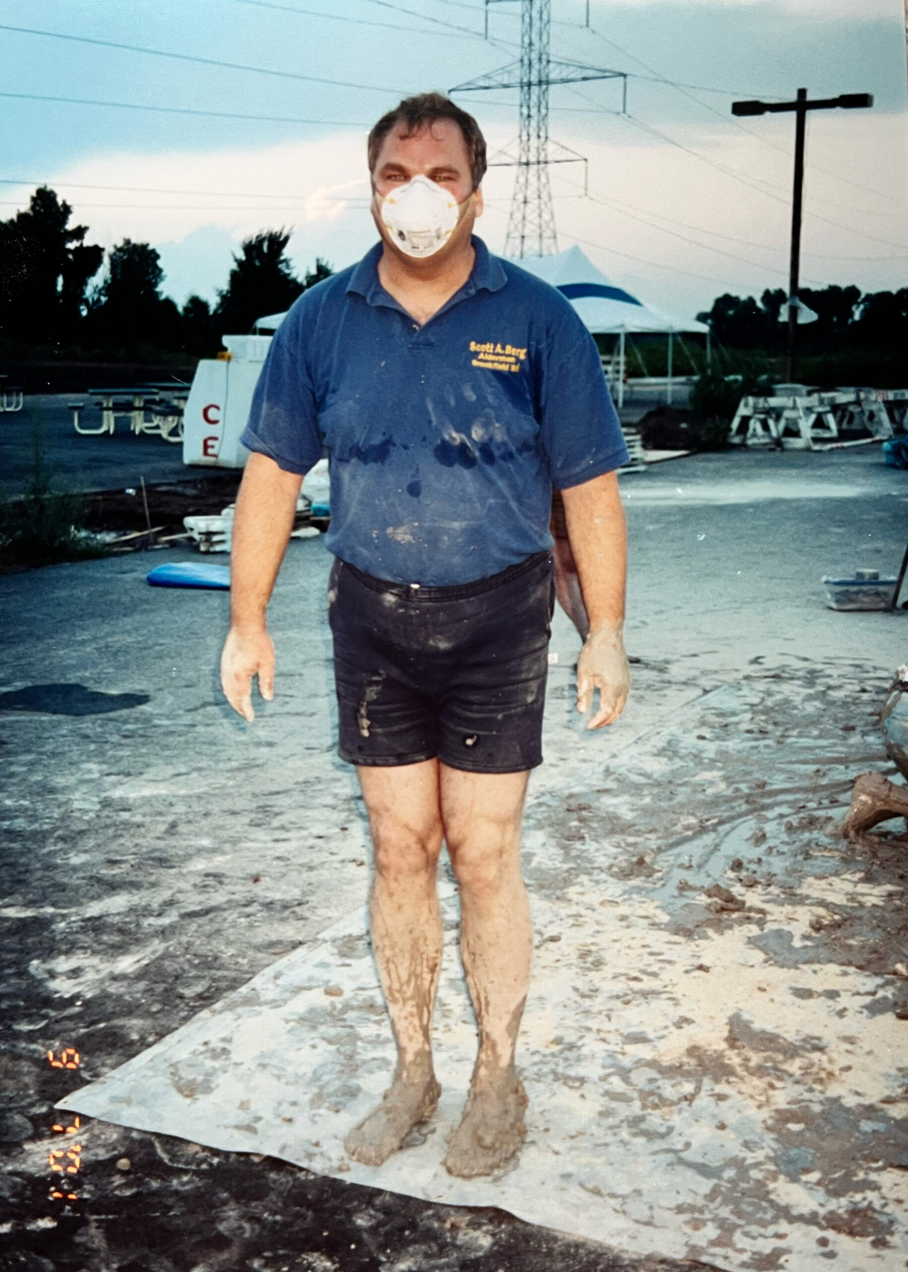 Man wearing a white protective mask and blue polo shirt stands on a muddy outdoor work site with power lines and tents in the background.