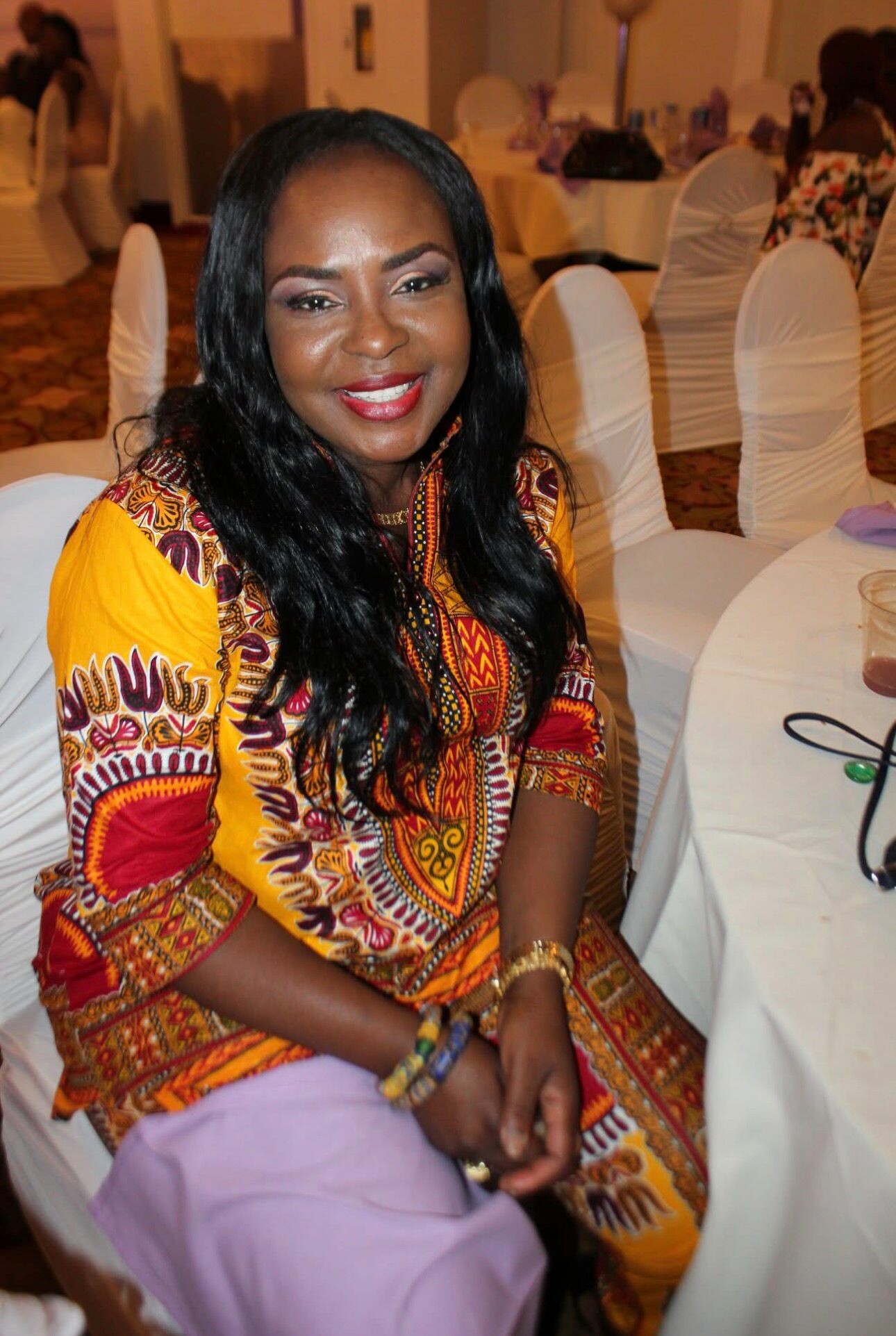 Smiling woman in a vibrant yellow and patterned dress sits at a banquet table in a formal event hall.