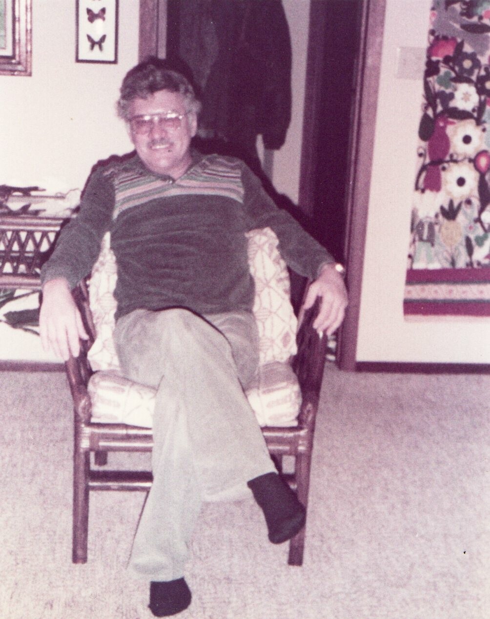 Older man with glasses lounging in a patterned armchair in a living room, legs crossed, wearing a striped sweater.