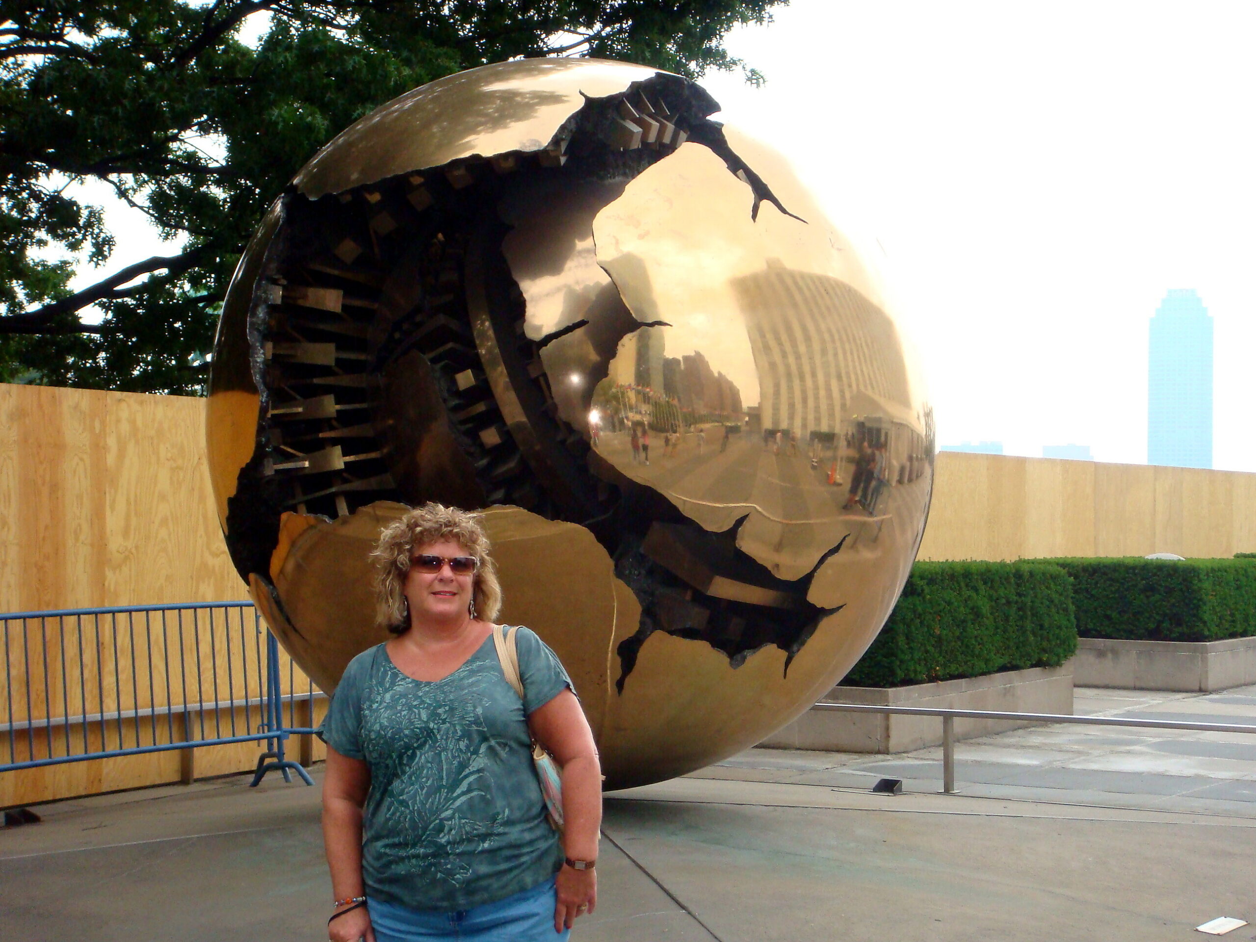 Woman in teal shirt with sunglasses standing in front of a cracked, reflective golden sphere sculpture outdoors.