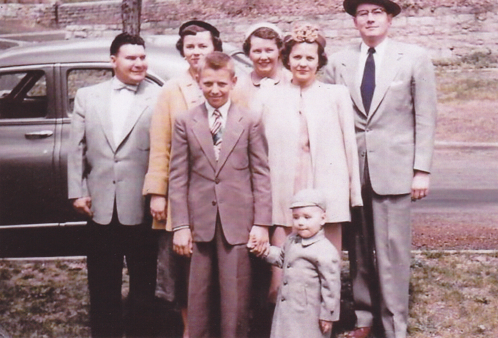 Family group portrait outdoors beside a dark car; adults in 1950s-style suits and dresses with a toddler in front holding hands.