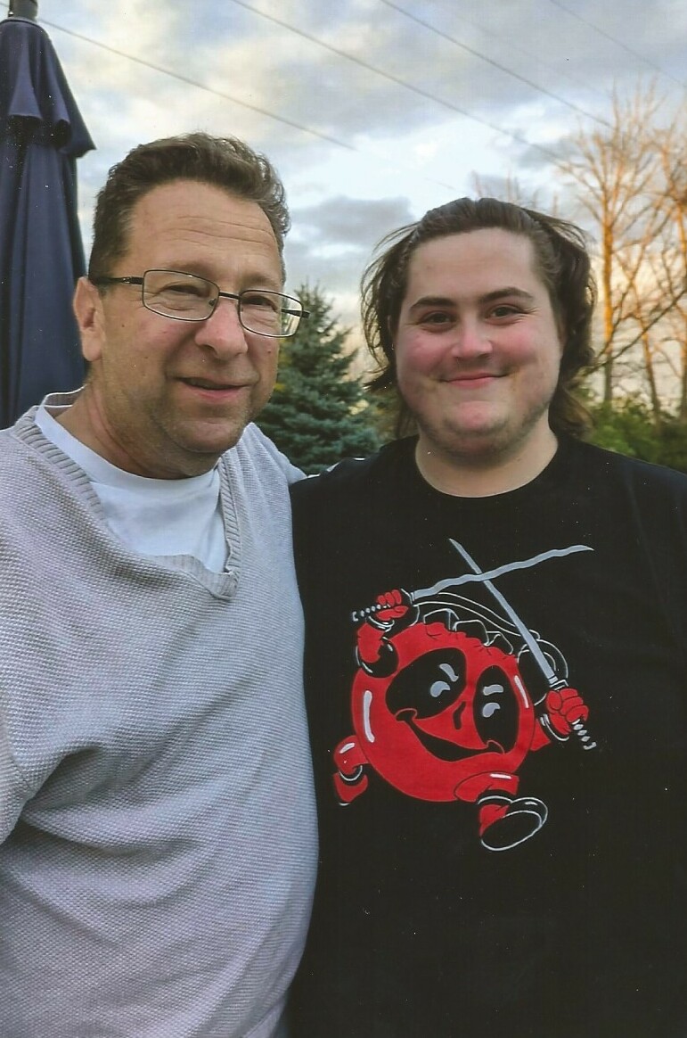Two adults pose together outdoors, smiling at the camera with trees and a blue sky in the background.