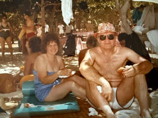 Couple relaxing on towels at a sunny outdoor gathering; the woman in a blue swimsuit sits beside the man in white shorts and a bandana, both smiling under a shaded area.