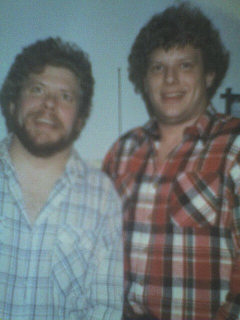Two men with curly hair and beards stand side by side, smiling at the camera, both wearing plaid shirts.