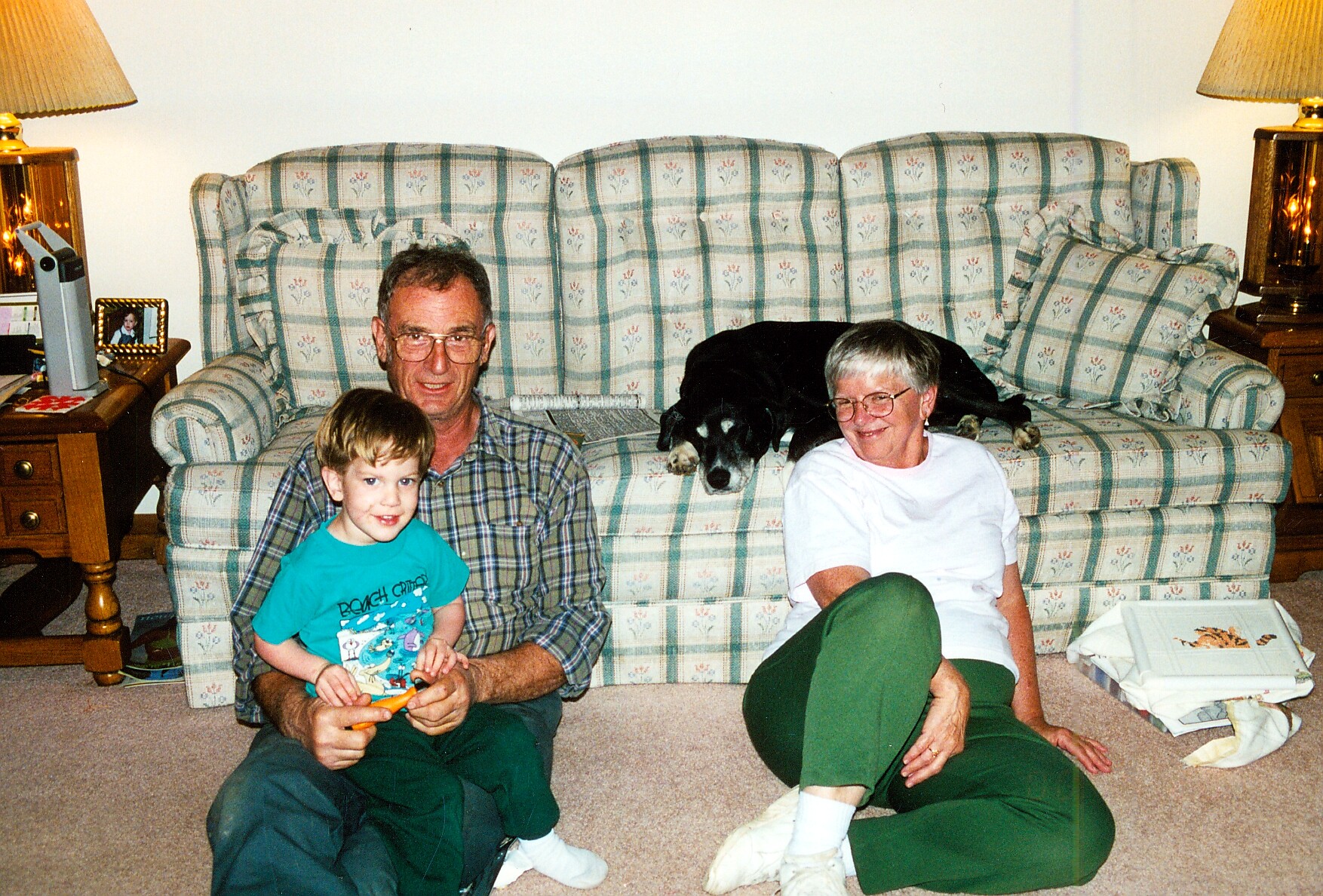 A man, a woman, and a small boy sit on a patterned sofa with a black dog resting between them in a living room.
