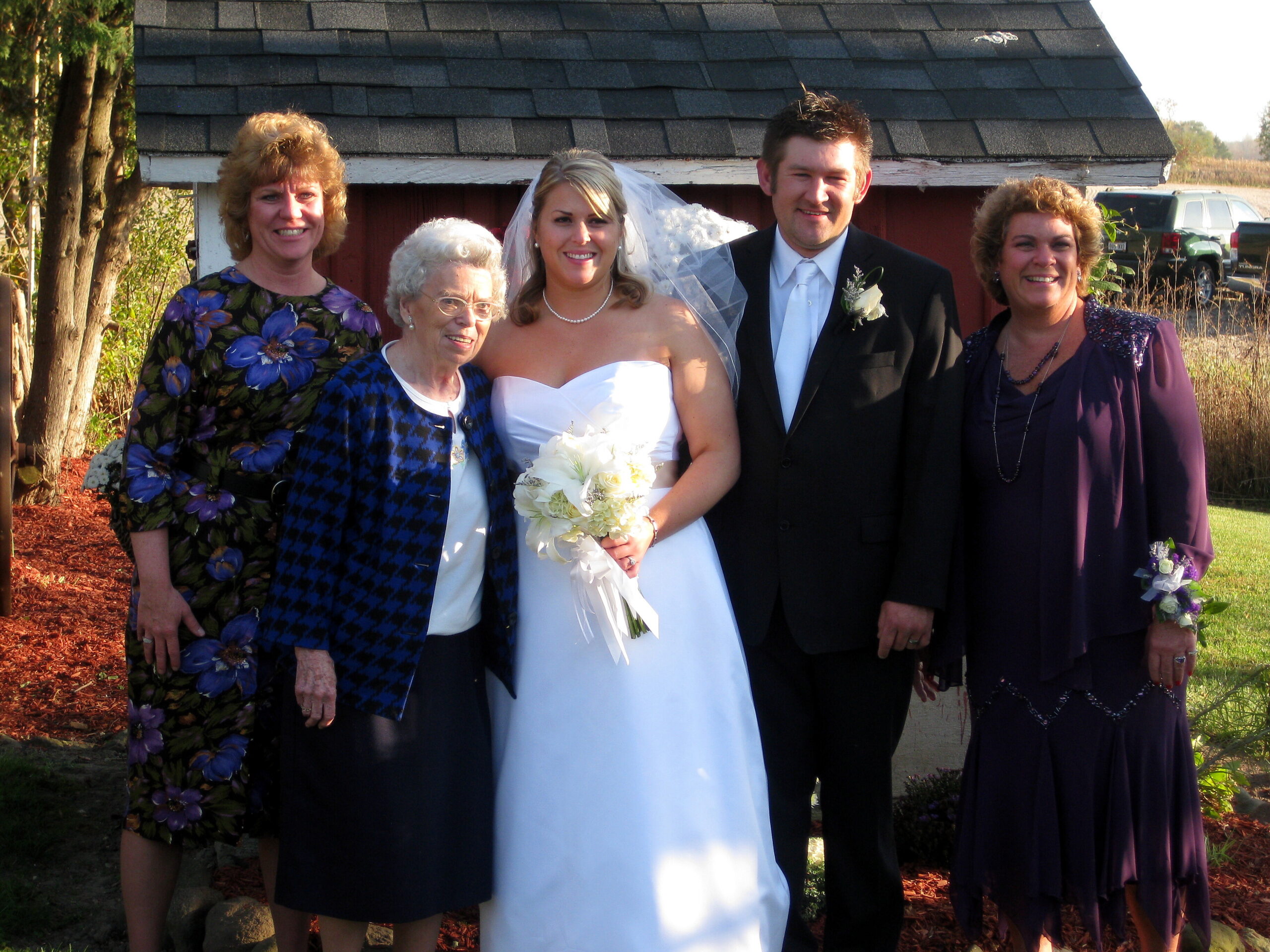 Bride in a white wedding dress holding a bouquet, standing with the groom and three relatives outdoors in front of a shed.