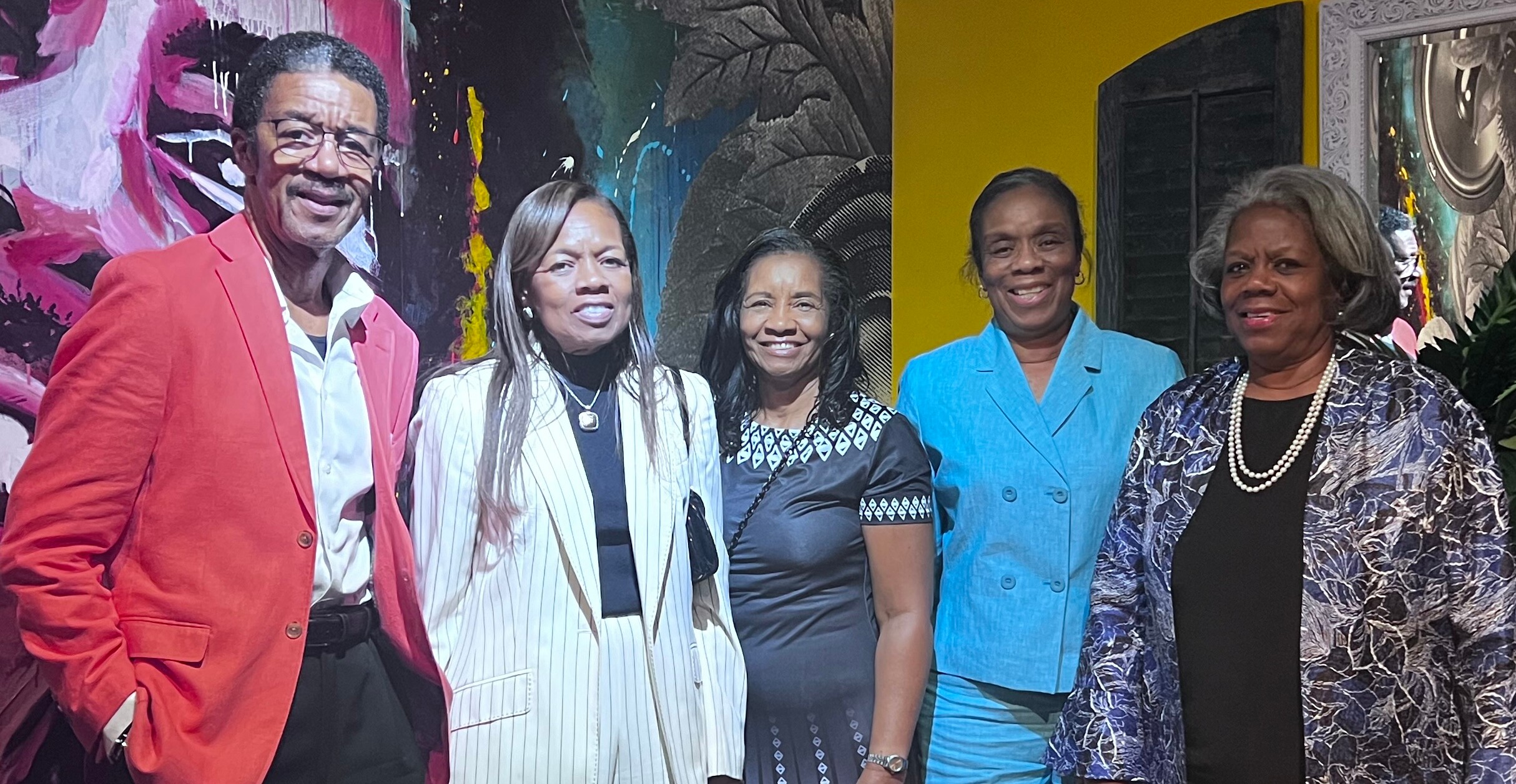 Group of five diverse adults posing and smiling in front of a colorful mural. (Left to right: man in red jacket, two women in light jackets, another woman in patterned dress, and a woman in a blue suit.)