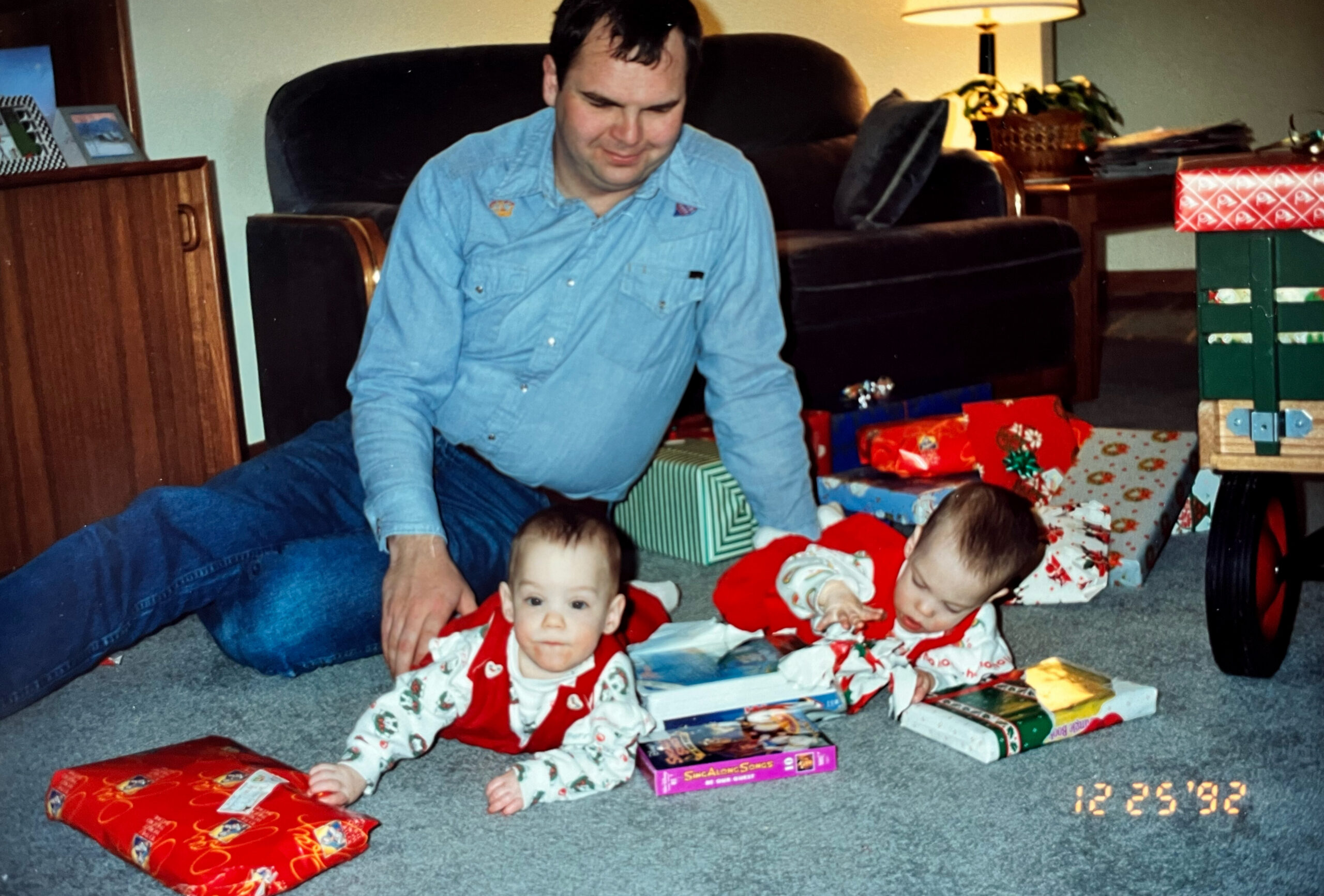 Man in a blue shirt sits on the carpet with two babies in festive outfits, surrounded by wrapped Christmas gifts in a living room.