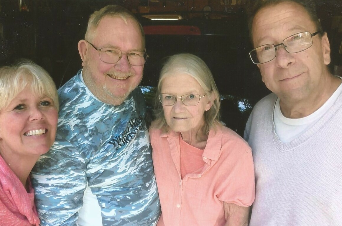 Four smiling adults posing closely for a group selfie outdoors, wearing casual shirts and sweaters with a pink blouse and blue patterned shirt visible in the scene.