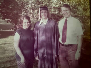 Young graduate in cap and gown stands between smiling adults outdoors, posing at a park on graduation day.