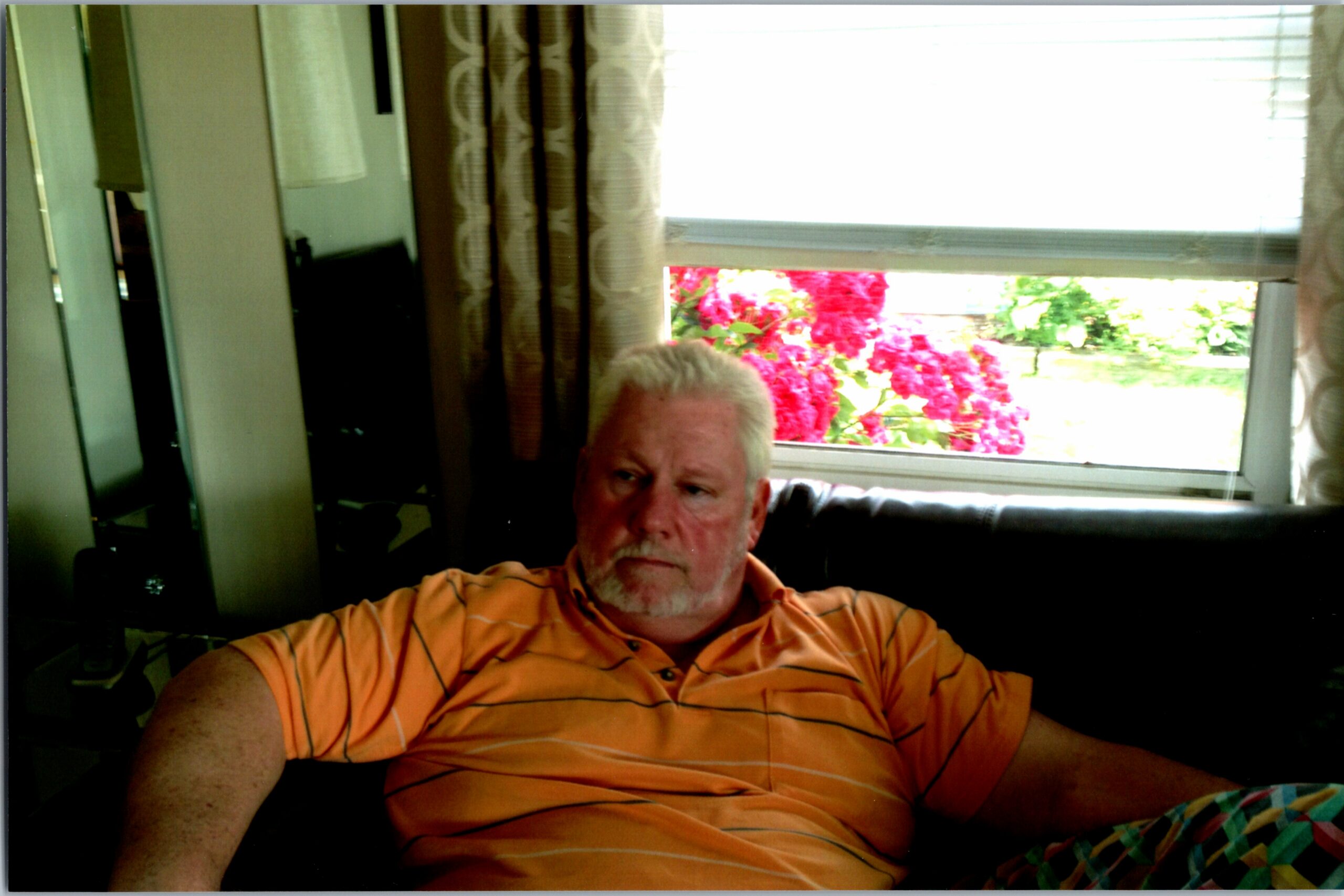 Older man with a white beard lounging on a dark sofa indoors, wearing an orange striped polo; window behind shows bright pink flowers outside.