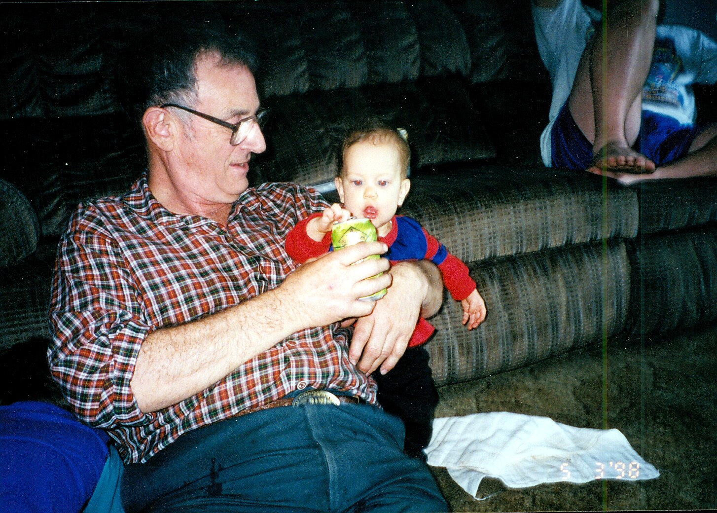 Older man in a plaid shirt sits on the floor with a baby on his lap, the baby reaching for a can held by the man in a living room setting.