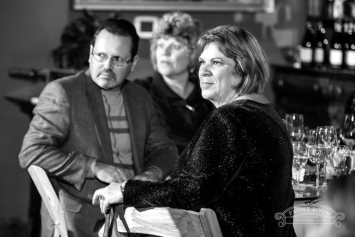 Three adults sit at a restaurant table; a woman in a glittery black top is foreground, with a man in a blazer to her left and a blurred woman in the background.