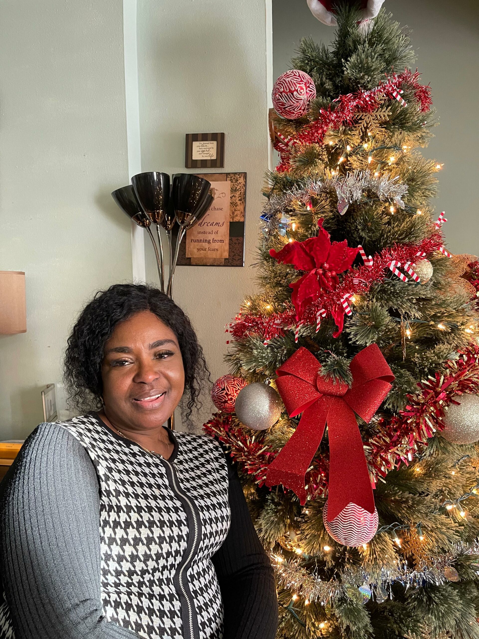 Smiling woman posing next to a decorated Christmas tree with red ribbons and lights.
