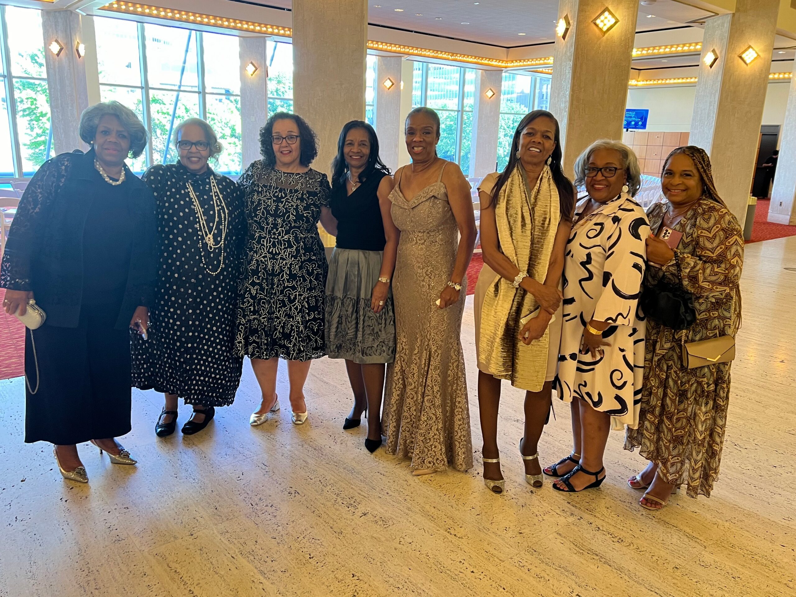 Group of seven women in formal dresses posing together indoors at a event venue or lobby.