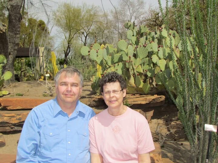 Two people sit on a wooden bench in a desert-style garden with cacti and succulents in the background.