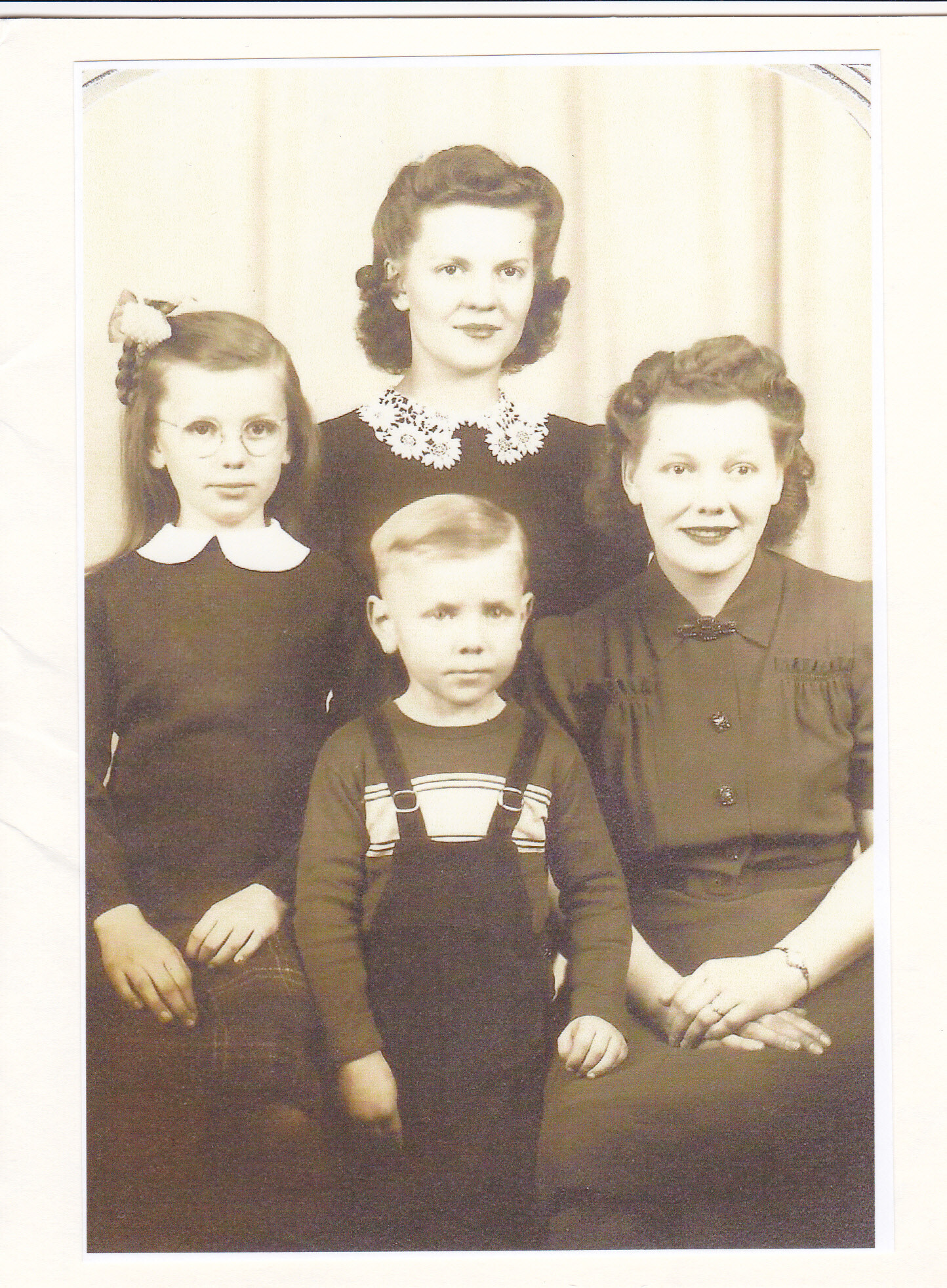 Family portrait of two women, a girl with glasses, and a small boy in vintage clothing posed together indoors.
