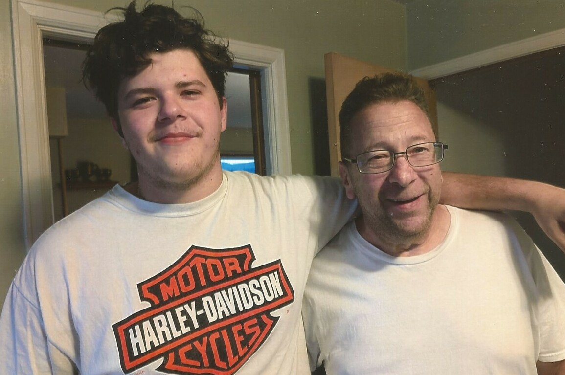 Two men stand close together indoors with their arms around each other, smiling at the camera (one wearing a Harley‑Davidson shirt).
