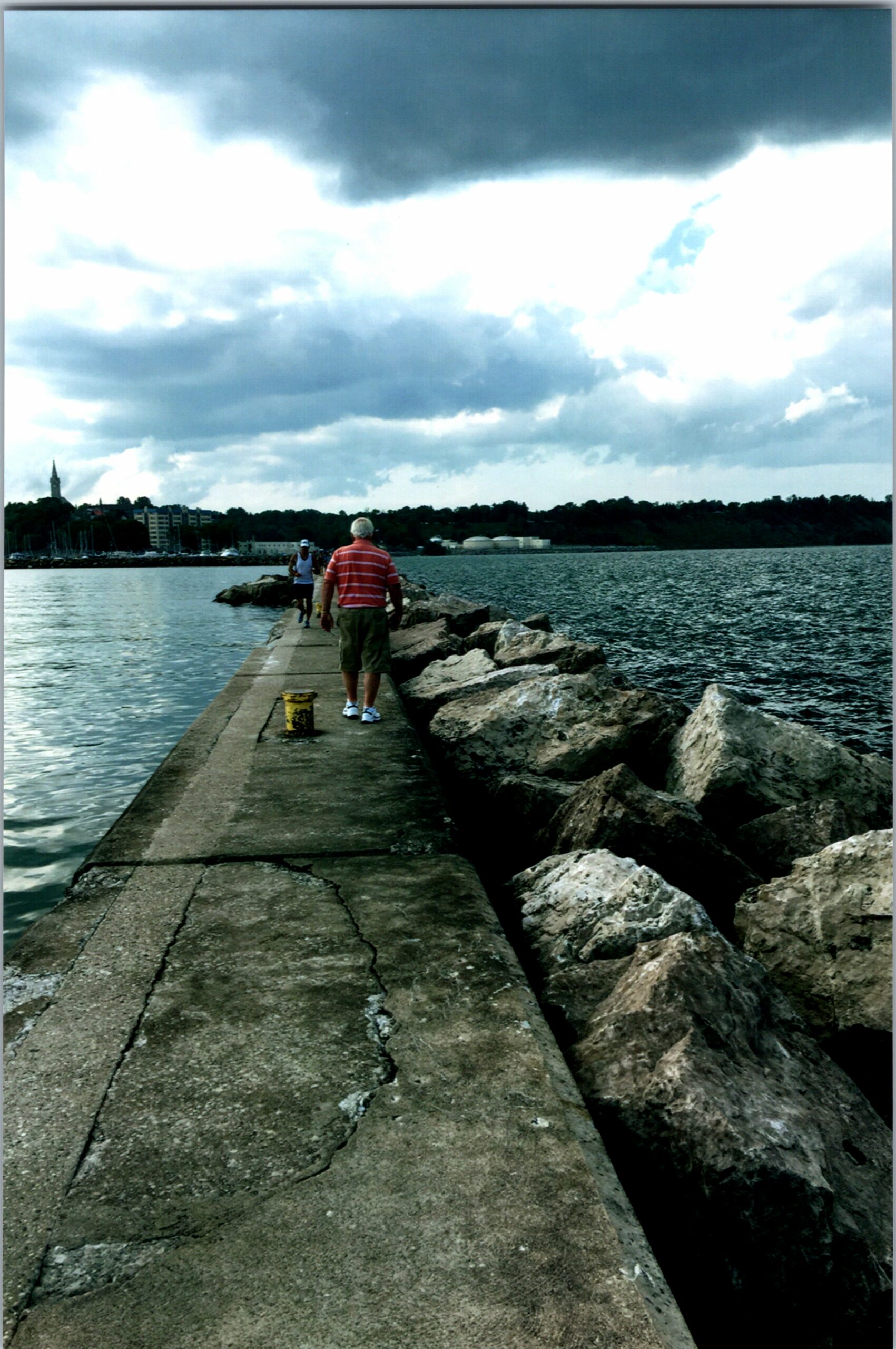 Two people walk along a concrete jetty that extends into a calm body of water, with large rocks on the right and a cloudy sky above.