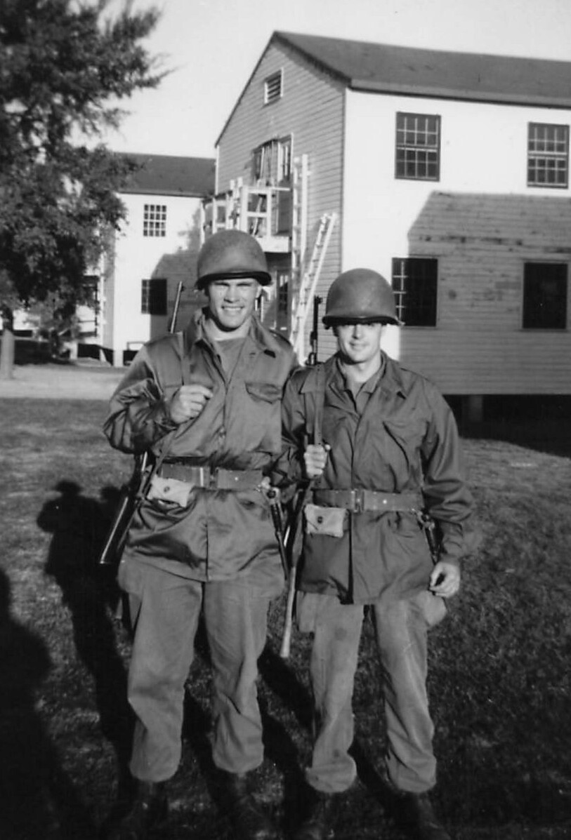 Two soldiers in WWII-era uniforms with helmets, standing side by side outside a wooden building, rifles slung over their shoulders.