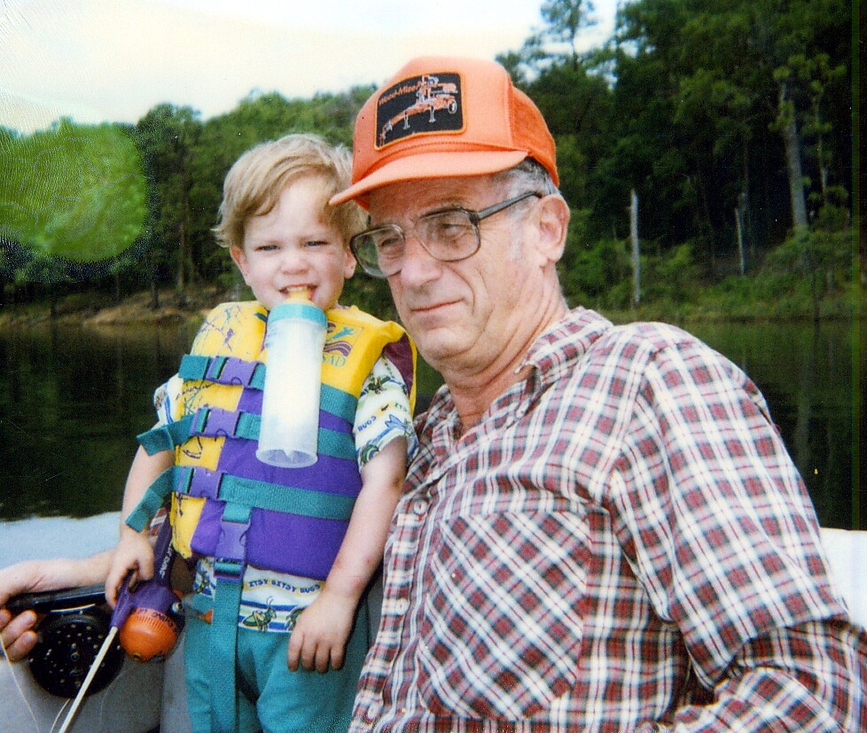 Grandfather in an orange cap and glasses sits next to a smiling toddler in a colorful life jacket on a boat by a calm lake.