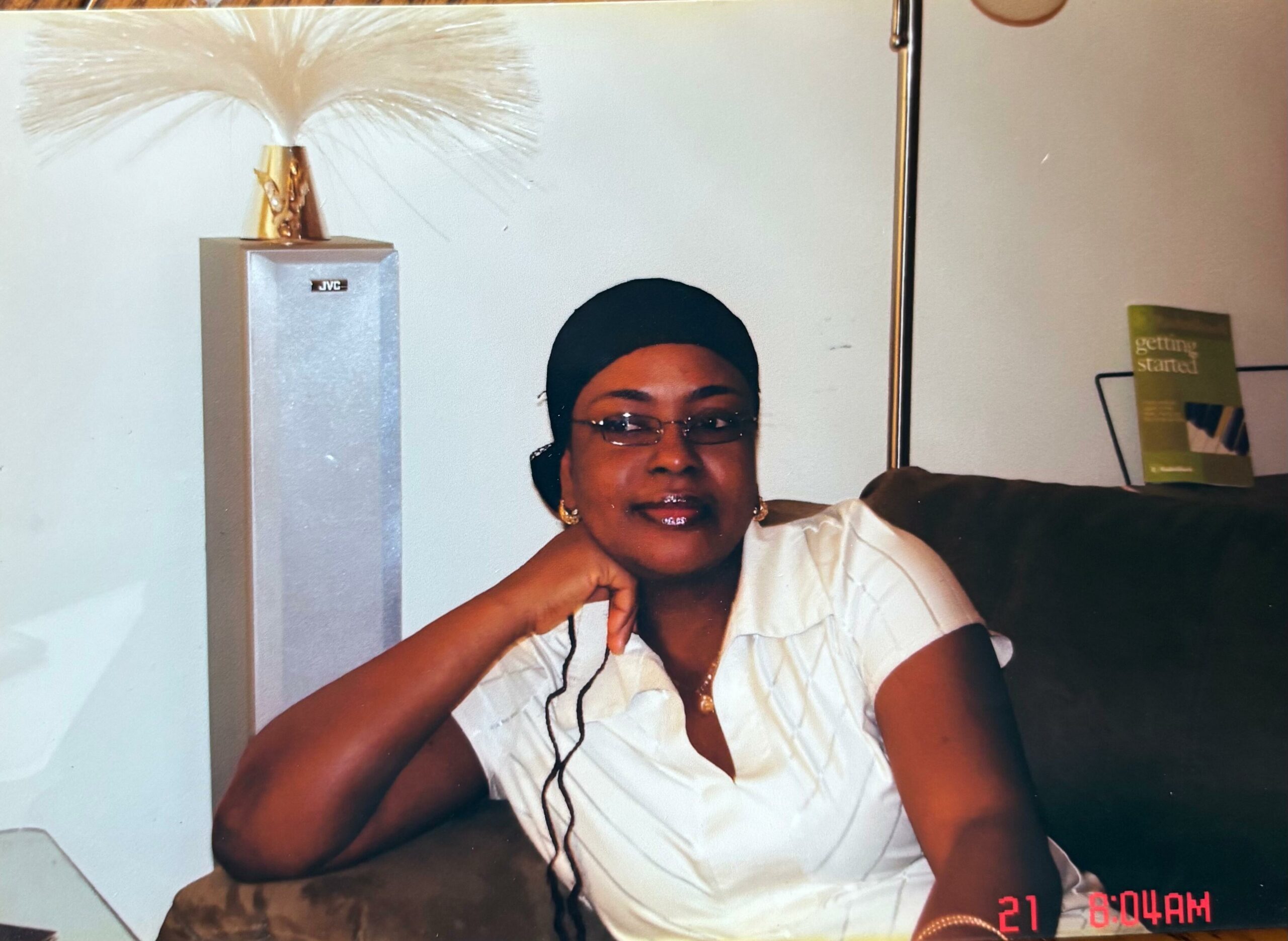 Portrait of a woman in a white blouse with a black head wrap, resting her chin on her hand, indoors with a feathered decor and a JVC speaker in the background.