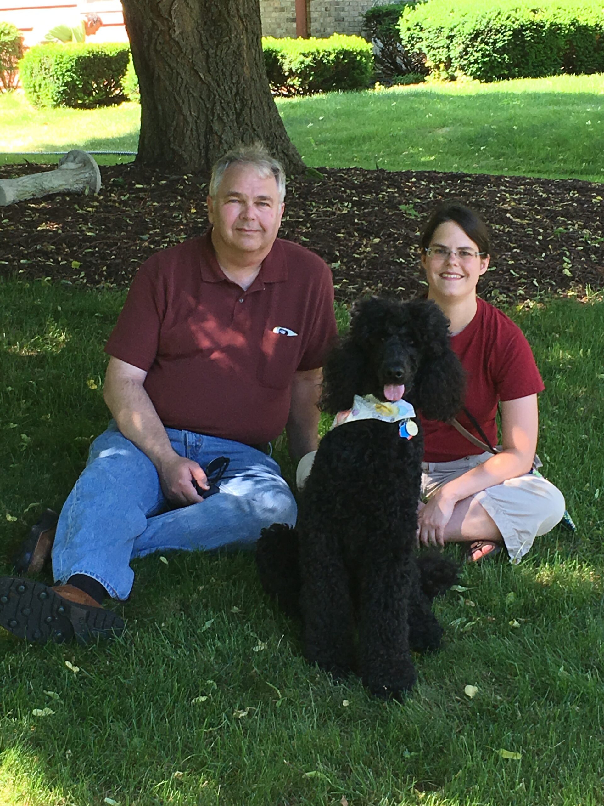 Couple sitting on grass under a tree with a black poodle wearing a scarf, in a park of sunny day.