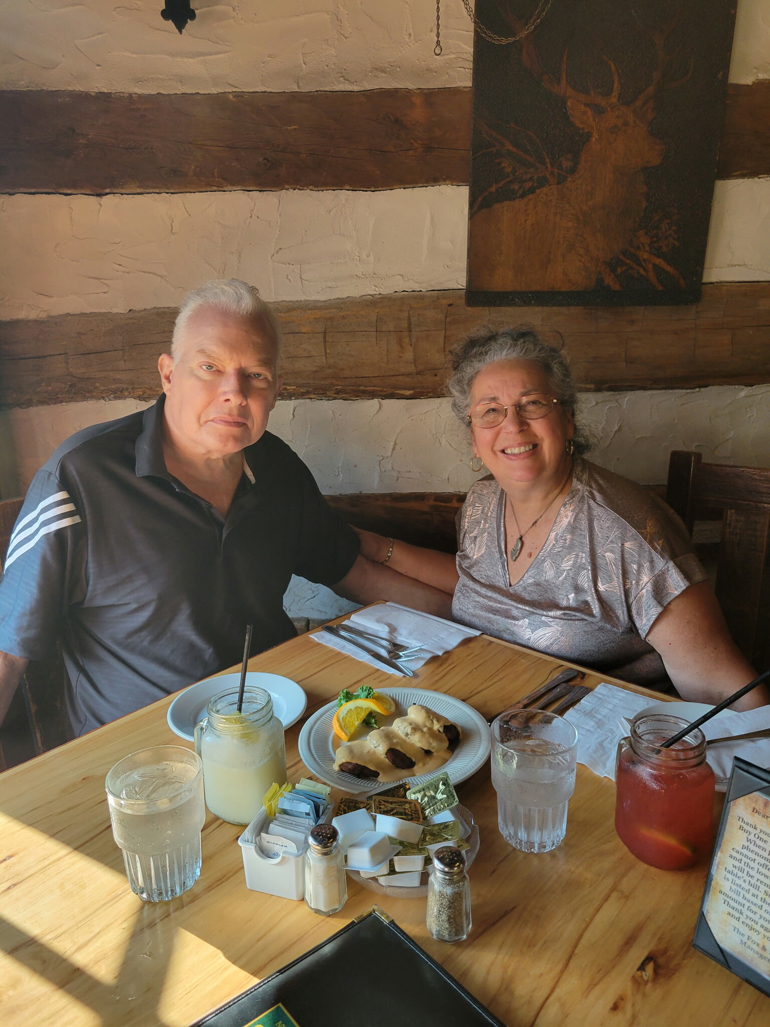 Two adults seated at a rustic wooden table in a restaurant, smiling at the camera with plates of food and drinks in front of them.