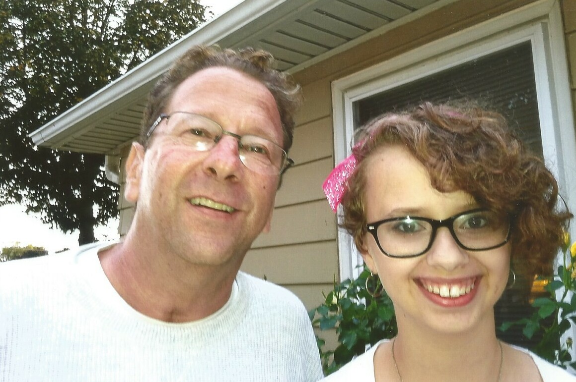 Two people smiling at the camera while taking a selfie outside a house in daylight.