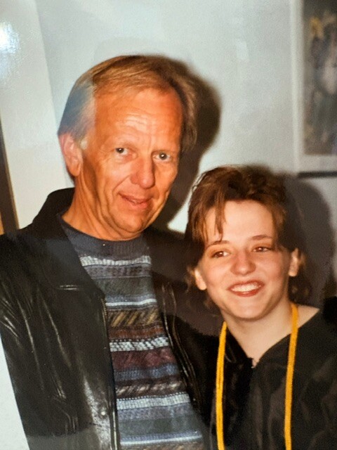 An older man in a leather jacket and a patterned sweater poses with a smiling younger woman indoors.