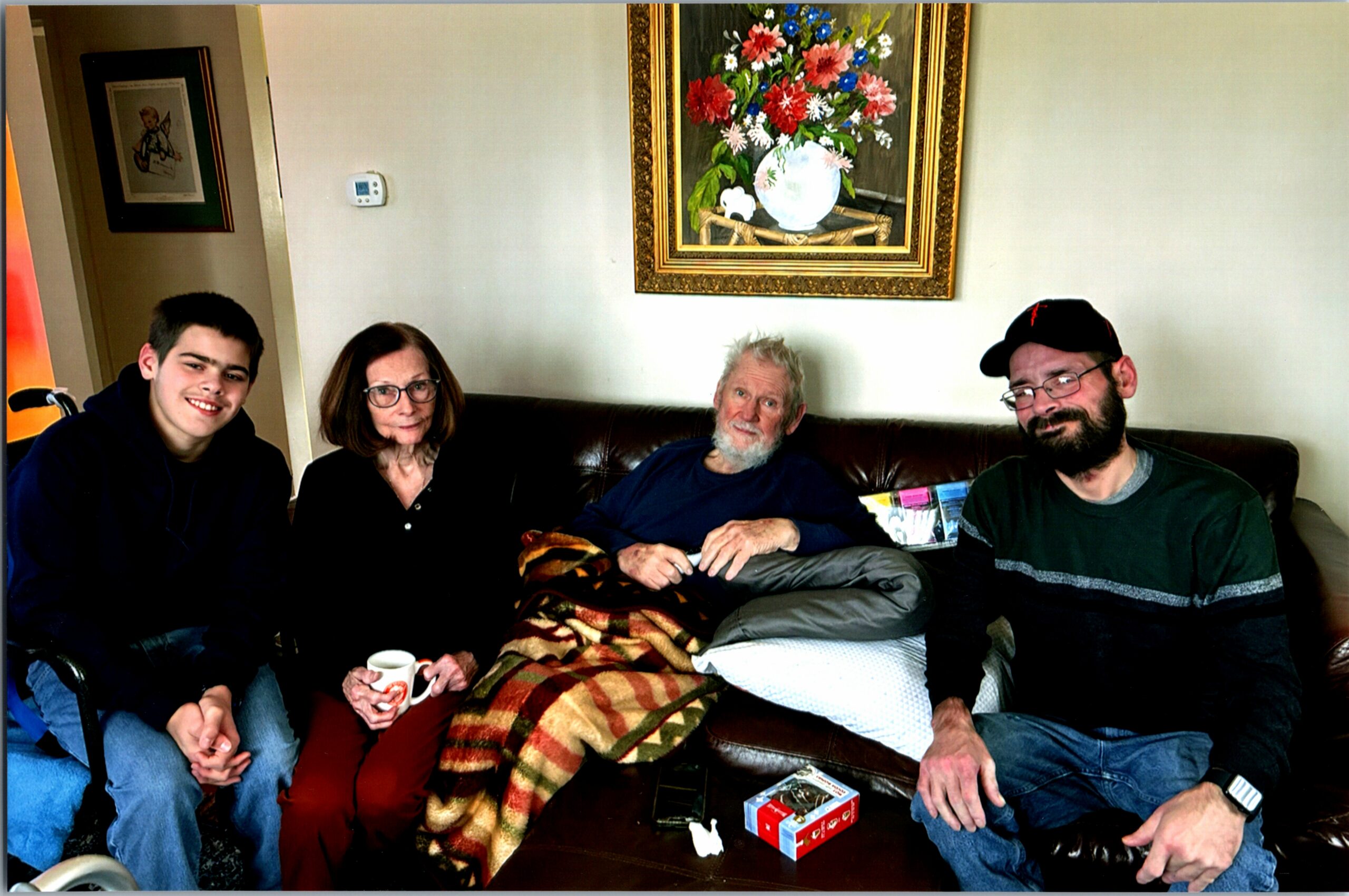 Four people sit on a brown leather couch in a living room, smiling at the camera with a striped blanket draped over their legs.