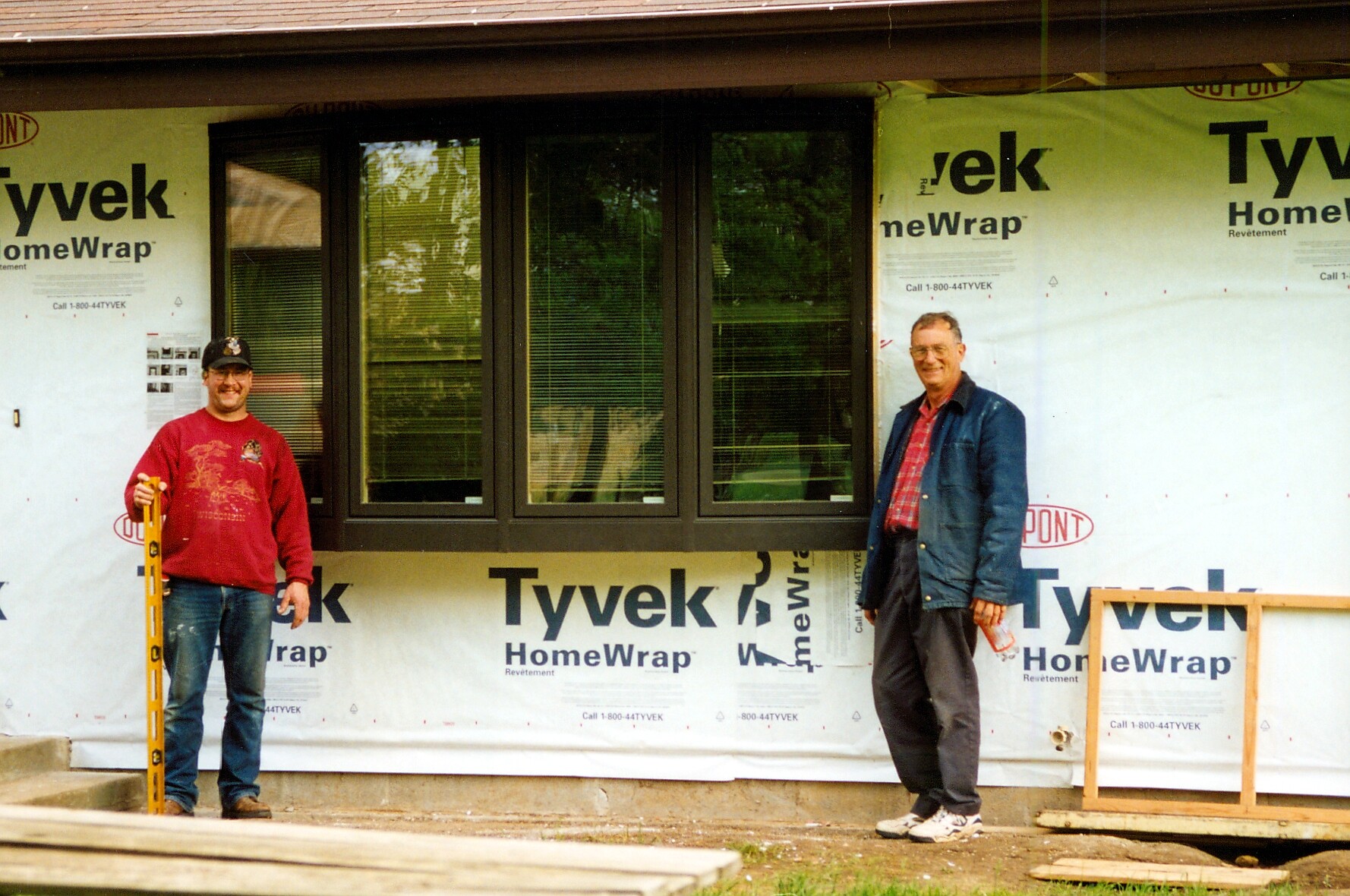 Two construction workers stand in front of a house under construction with a large triple window and Tyvek wrap behind them.