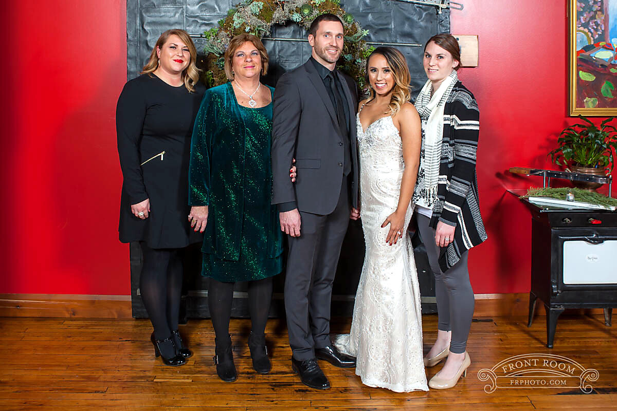 Bride in a white gown and groom in a gray suit pose with three guests indoors against a red wall and wooden floor.