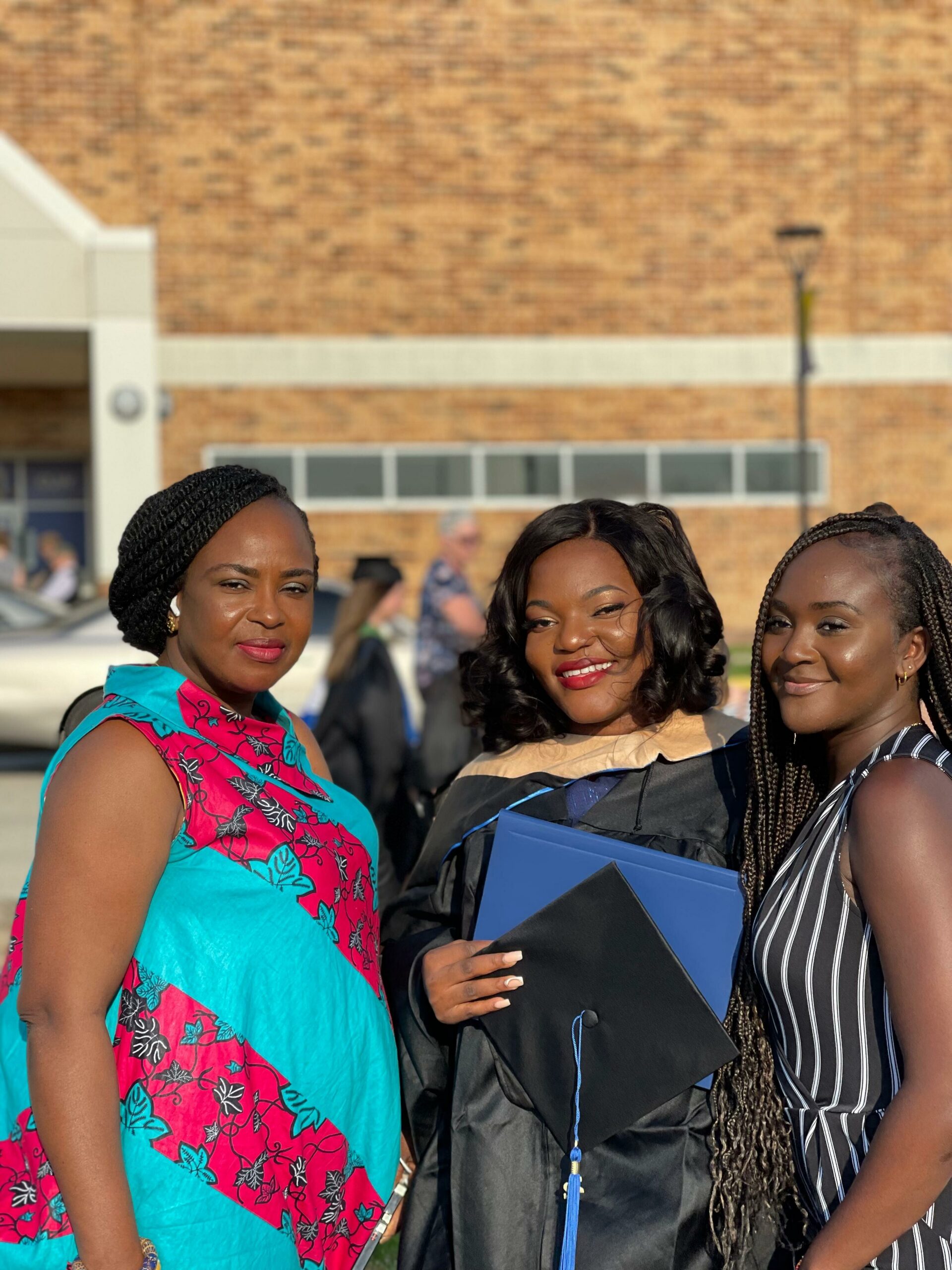 Three smiling women posing outdoors at a graduation, one in a colorful dress, another in a gown with a blue diploma folder, and the third in a striped dress with long braids.