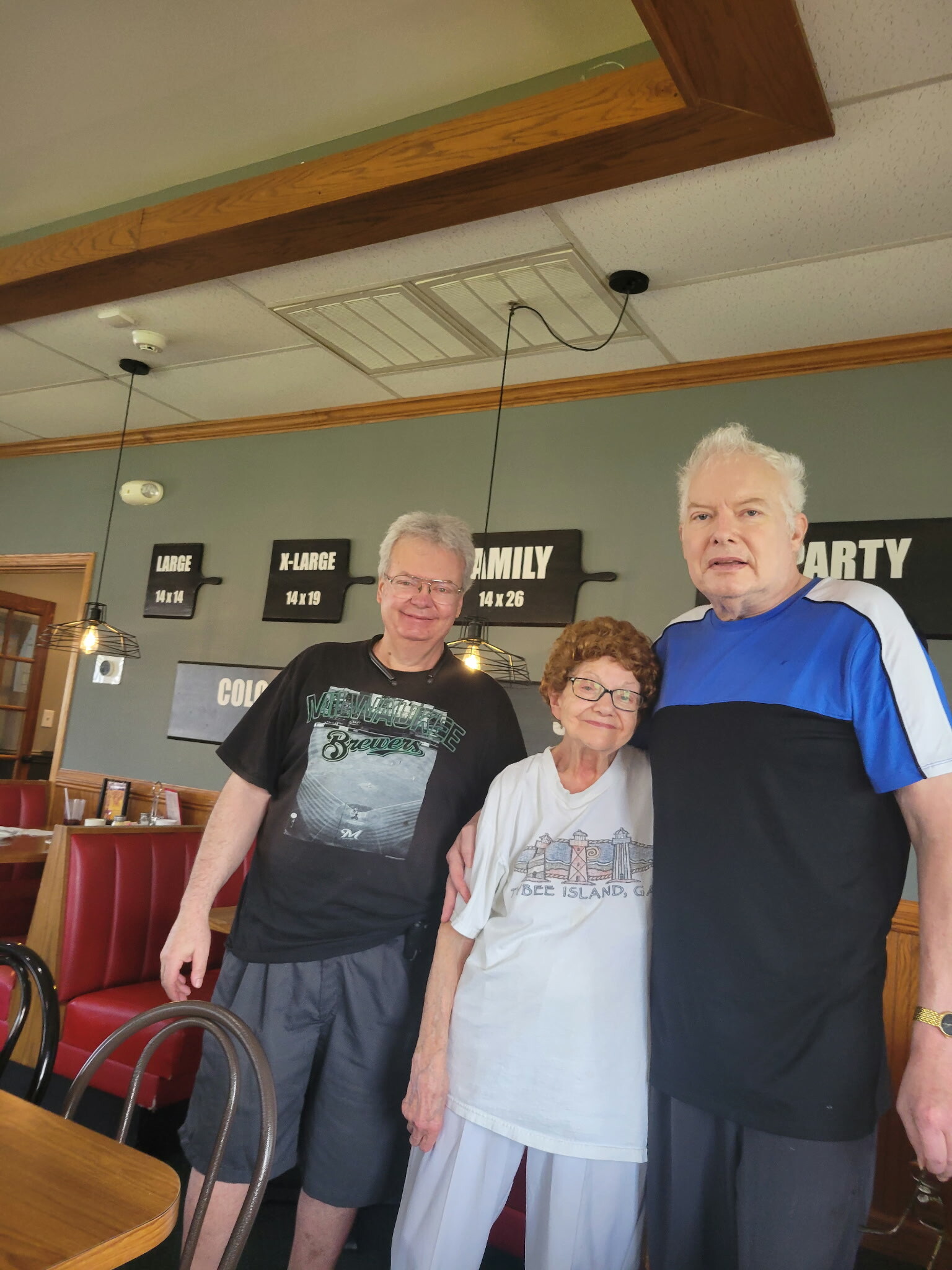 Three smiling seniors posing together in a casual restaurant, with signs and ceiling fixtures in the background.