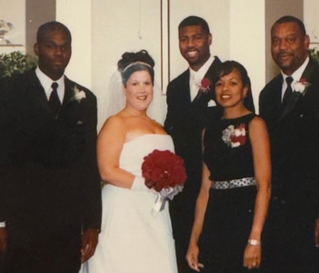 Bride in a white gown with veil, holding a red bouquet, posing with the groom and two guests in formal attire at a wedding.