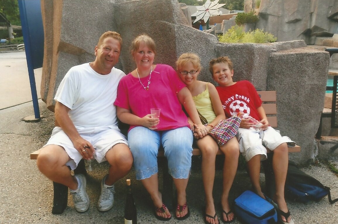 A family of four sits on a park bench posing for a photo; man in white shirt, woman in pink, two kids smile beside her with drinks in hand.