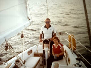Man stands at the boat's helm while a woman sits in the cockpit on a sailboat cruising on calm water, sunny day.