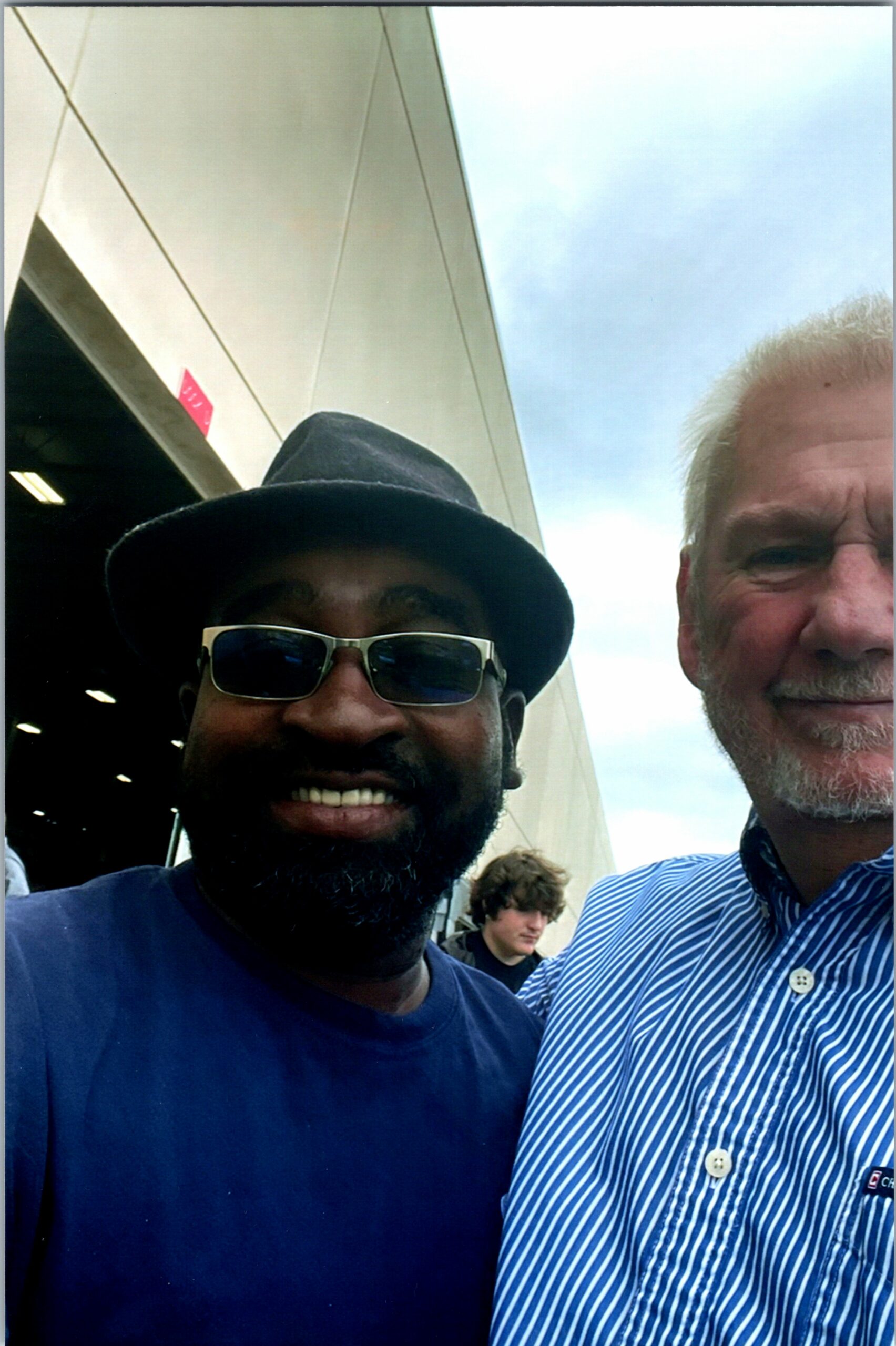 Close-up selfie of two men outdoors: man on left wearing a black fedora and sunglasses, man on right in a blue-striped shirt.