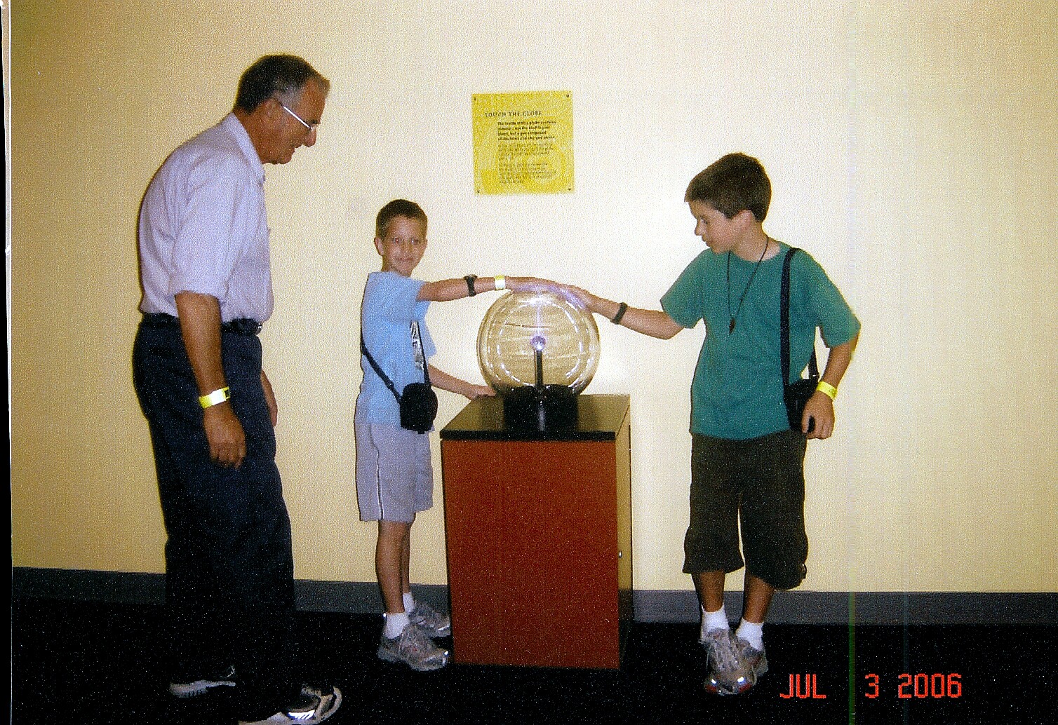 Older man and two boys touch a glowing plasma globe on a pedestal in a science museum.