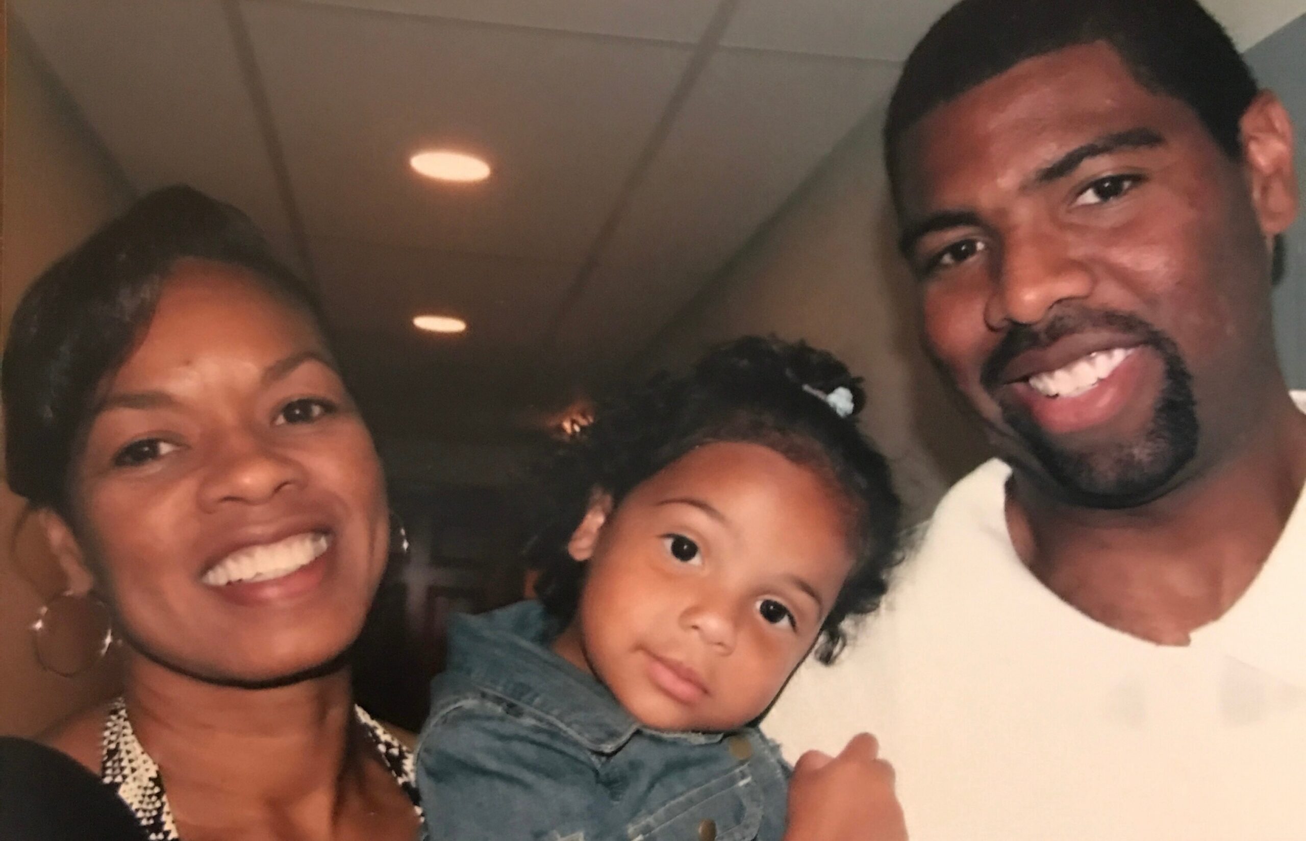 Smiling woman on the left, a young child in the center, and a smiling man on the right indoors in a close family photo.